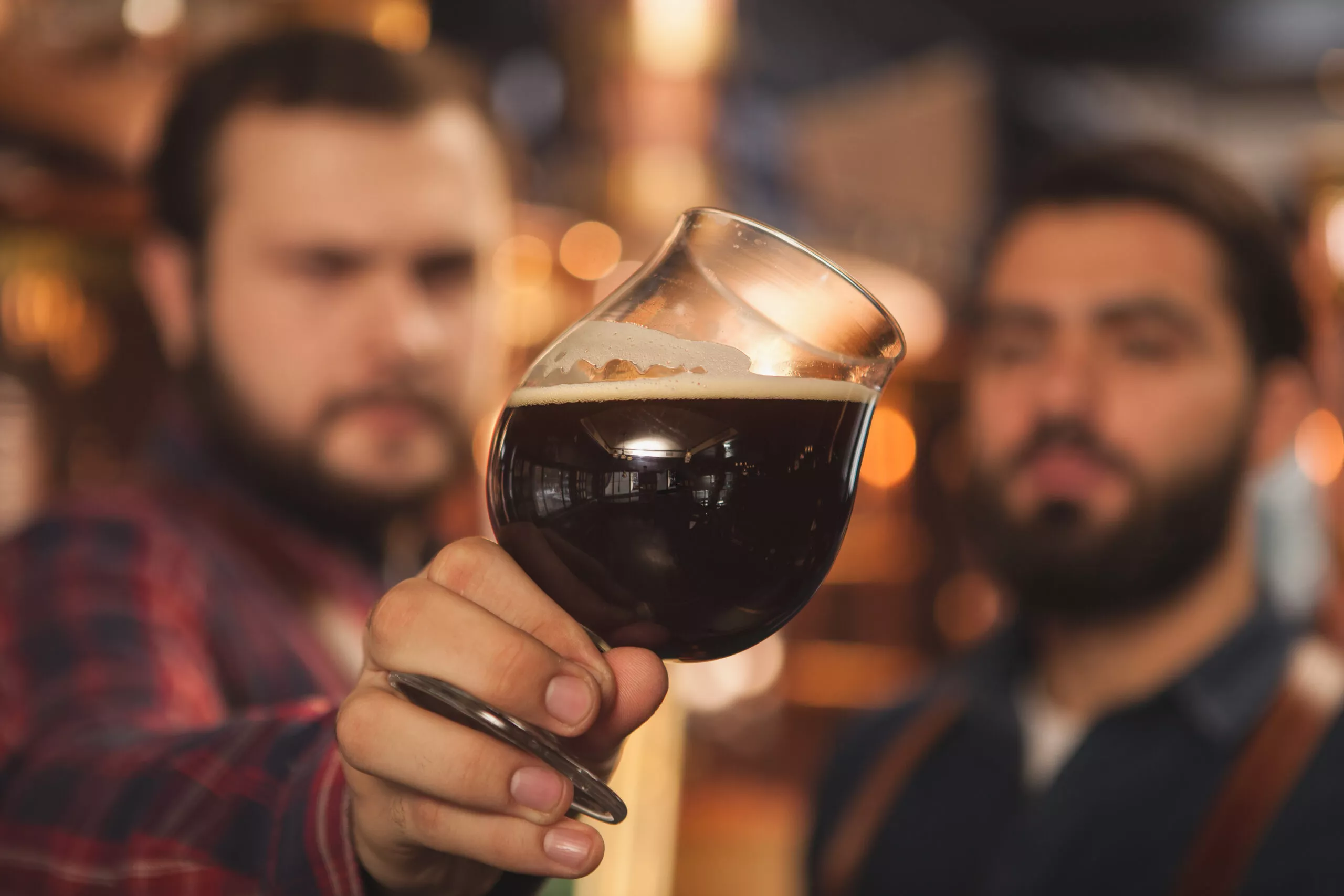 Close up shot of two brewers examining delicious dark beer in a glass, working at the beer production factory. Selective focus on a beer mug in the hands of beermakers. Quality, expertise concept