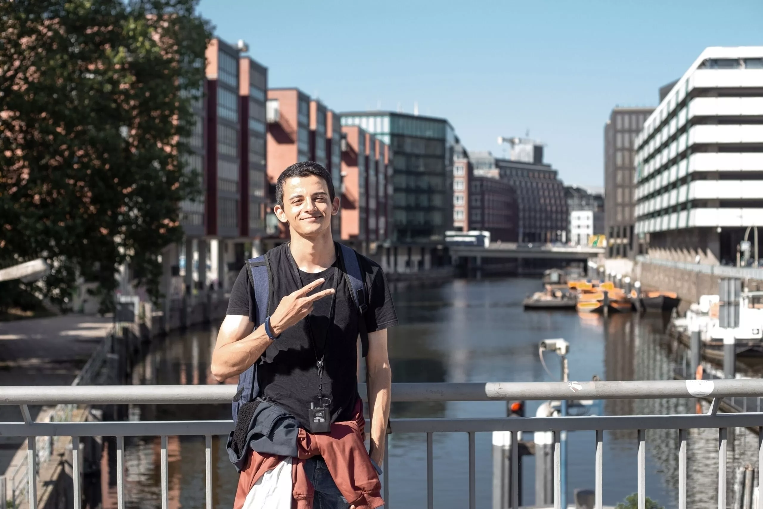 Молодой мужчина со средним востоком и рюкзаком во время экскурсии по центру Гамбурга в Германии с помощью аудиогида и фотографируется на фоне городских каналов в солнечный летний день.
