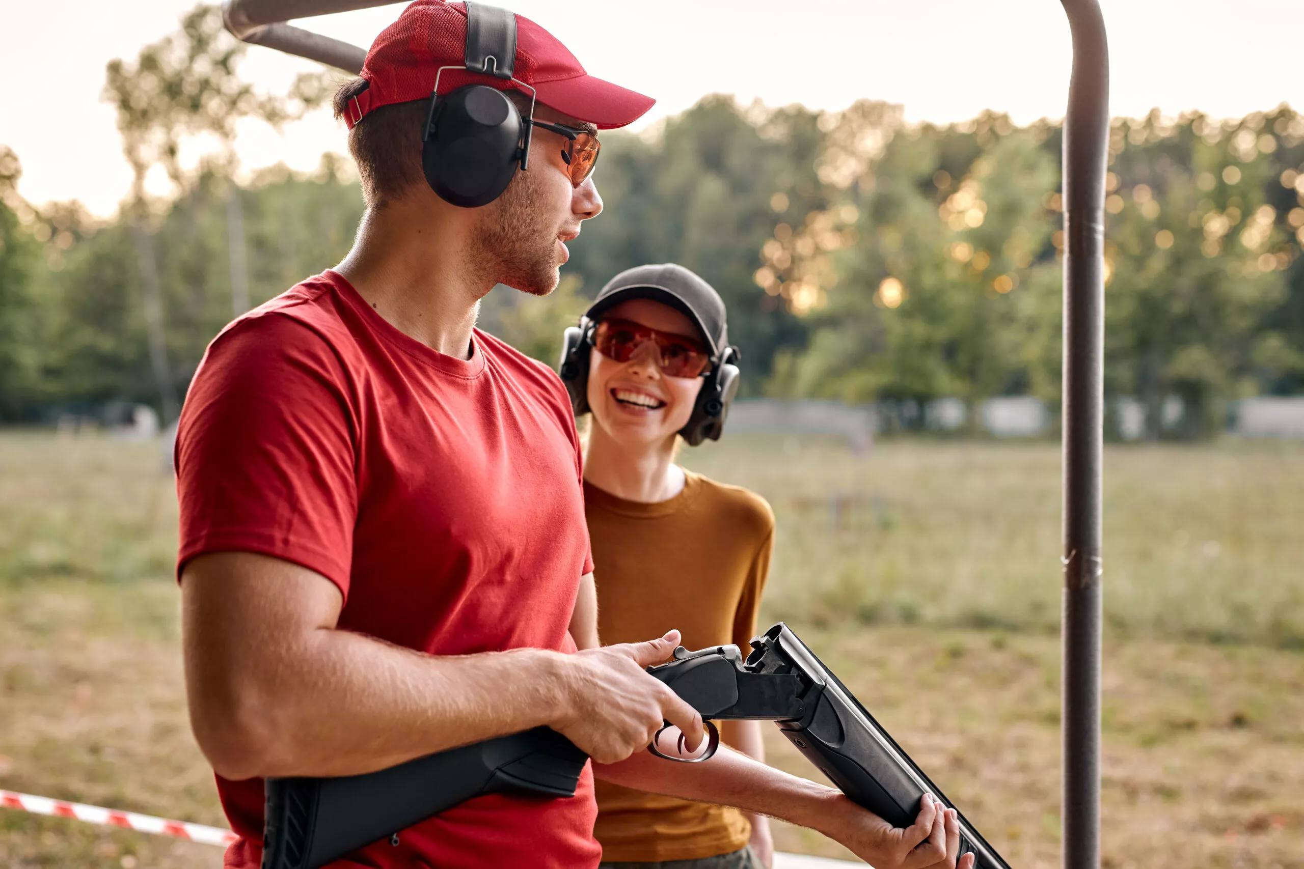 Handsome male shooting instructor teaches the backslider how to handle weapon on training course in outdoor range. cheerful teacher and woman in protective spectacles and headset have fun, laugh